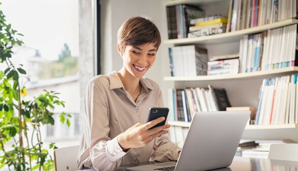Frau sitzt lachend am Schreibtisch mit einem Smartphone in der Hand. Vor ihr steht ein Notebook.