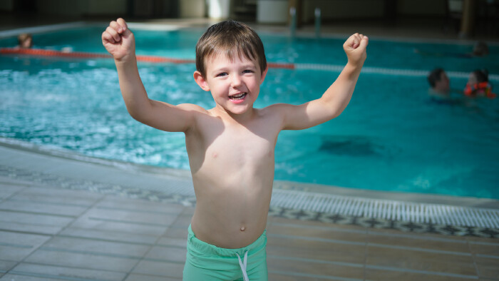 Fröhlicher Bub im Hallenbad © Андрей Журавлев, stock.adobe.com Fröhlicher, der Kamera zugewandter Bub mit gehobenen Armen steht vor einem Pool im Hallenbad.