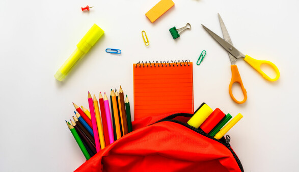 Bunte Schulmaterialien auf wei&szlig;em Hintergrund, darunter Stifte, Notizbuch, Schere und ein roter Rucksack.
