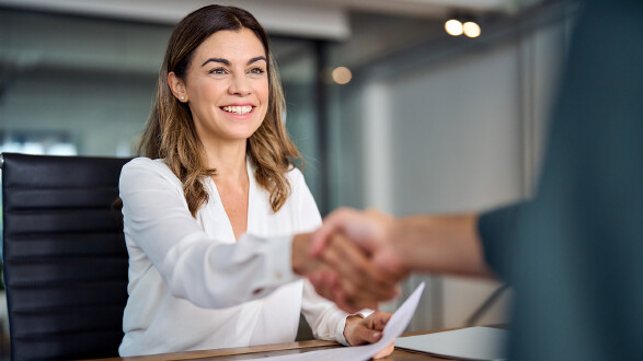 Eine Frau sitzt an einem Tisch in einem B&uuml;ro und sch&uuml;ttelt einer anderen Person die Hand. In der Hand h&auml;lt sie ein Dokument, vermutlich Bewerbungsunterlagen. Die Szene zeigt ein Bewerbungsgespr&auml;ch.