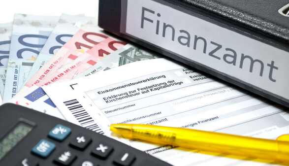Auf einem Tisch liegen Geld, ein Stift und ein Taschenrechner, eine Einkommensteuererklärung und ein Ordner mit der Aufschrift "Finanzamt"
