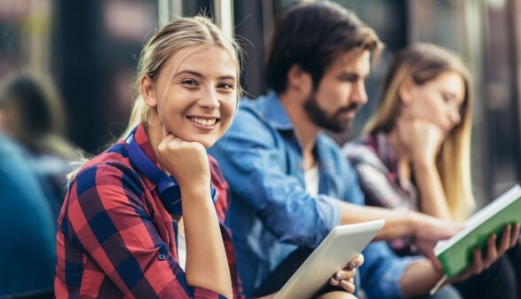 Junge Frau mit Tablet in der Hand l&auml;chelt in die Kamera, im Hintergrund unscharf sitzen ein weiterer Mann und eine Frau mit Unterlagen in der Hand