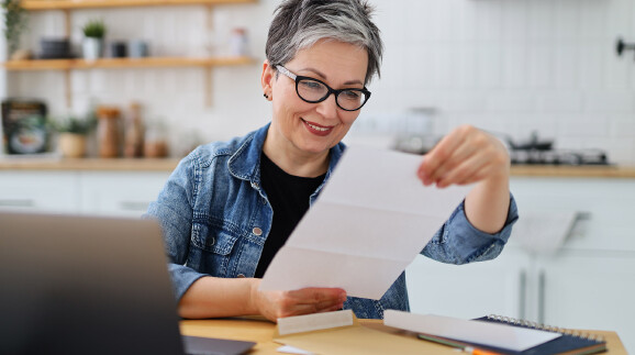 Eine Frau mit grauen Haaren und Brille sitzt in ihrer Küche und liest auf einem Blatt.