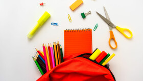 Bunte Schulmaterialien auf weißem Hintergrund, darunter Stifte, Notizbuch, Schere und ein roter Rucksack.